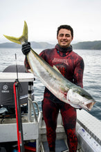 Load image into Gallery viewer, Man in a wetsuit holding a large fish on a boat with water and mountains in the background
