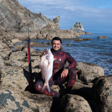 Load image into Gallery viewer, Man in a wetsuit holding a fish on rocky coastal terrain with ocean and cliffs in the background
